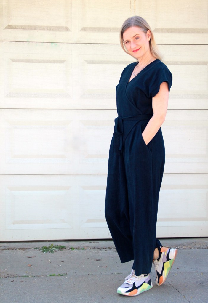 Frivolous at Last: Zadie Jumpsuit. Lori stands sideways to the camera with her hands in her pockets, smiling and wearing a navy blue jumpsuit that wraps at the front and has a tie belt, and colourful sneakers.