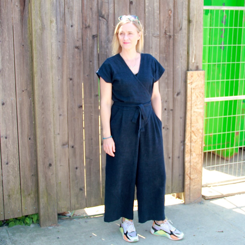 Frivolous at Last: Zadie Jumpsuit. Lori is standing in front of a wooden fence wearing a navy blue jumpsuit that wraps at the front and has a tie belt, and colourful sneakers.